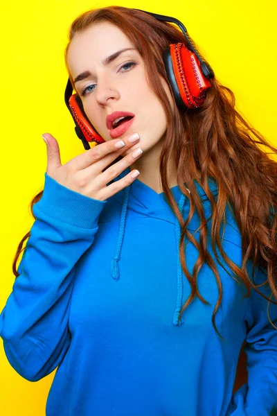 Молодая сексуальная девушка-диджей в синей куртке слушает музыку в красных наушниках на желтом фоне и веселится — стоковое фото