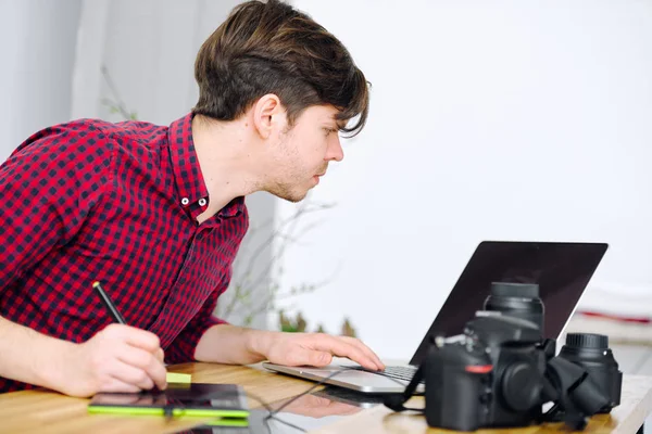 Молодий милий хлопець працює в офісі з ноутбуком і редагує фотографії за допомогою графічного планшета — стокове фото
