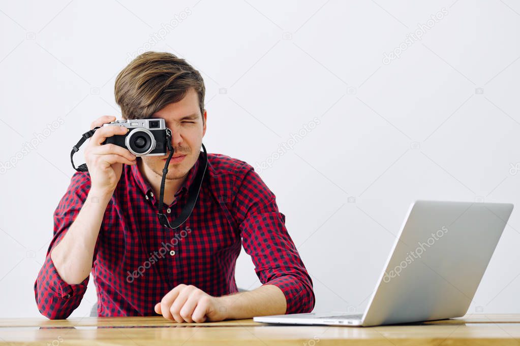 Young and successful man uses Mirrorless camera and sits at a table with a laptop in the office