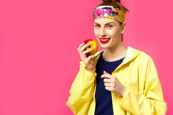 Giovane donna su uno sfondo rosa e un cappotto giallo tiene in mano una mela. Concetto di alimentazione sana e sport. Il concetto di ossessione per il colore. Stile minimalista. Trendy alla moda — Foto Stock