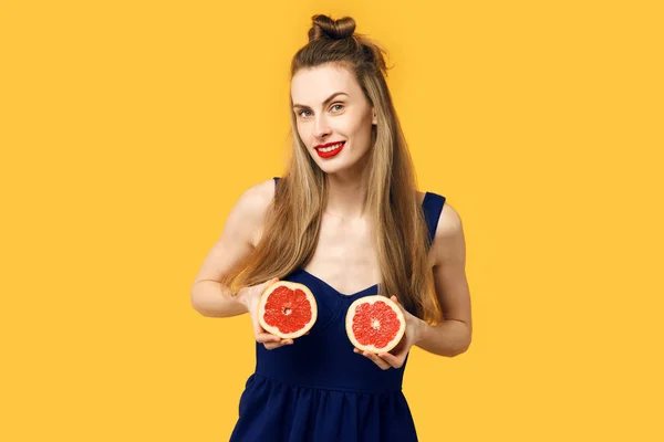 Mujer joven sobre un fondo amarillo y un vestido azul sostiene un corte naranja en sus manos. Concepto de nutrición saludable y deportes. Concepto de obsesión por el color. Estilo minimalista. Moda con estilo — Foto de Stock