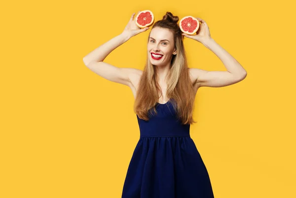 Mujer joven sobre un fondo amarillo y un vestido azul sostiene un corte naranja en sus manos. Concepto de nutrición saludable y deportes. Concepto de obsesión por el color. Estilo minimalista. Moda con estilo — Foto de Stock