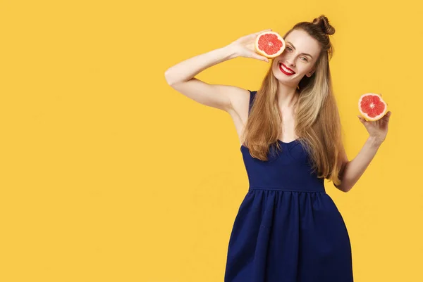 Mujer joven sobre un fondo amarillo y un vestido azul sostiene un corte naranja en sus manos. Concepto de nutrición saludable y deportes. Concepto de obsesión por el color. Estilo minimalista. Moda con estilo — Foto de Stock