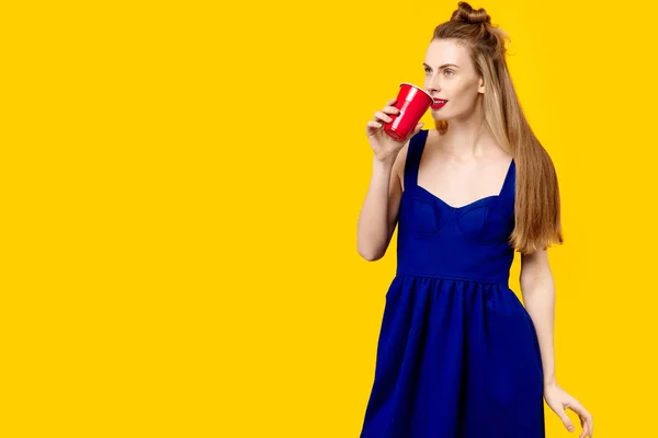 Giovane donna su uno sfondo giallo e un vestito blu sta tenendo un bicchiere. Il concetto di ossessione per il colore. Stile minimalista. Trendy alla moda — Foto Stock