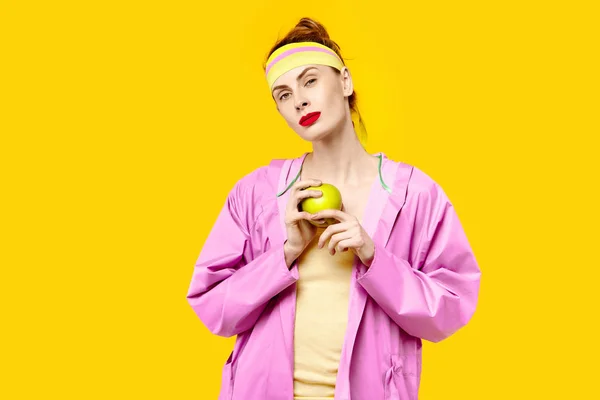 黄色背景和粉红色大衣的年轻女子拿着一个苹果。色彩痴迷的概念。简约风格。时尚时尚 — 图库照片