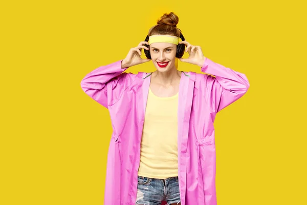 年轻女子在一个黄色的背景和粉红色的大衣拥抱音乐蠕虫无线耳机。色彩痴迷的概念。简约风格。时尚时尚 — 图库照片