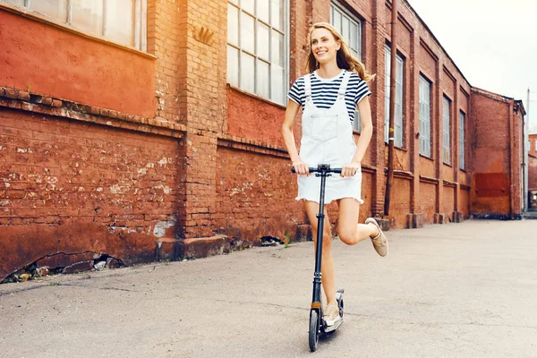 Menina bonita jovem está montando uma scooter no verão ao longo da rua na scooter e ri. O conceito de um estilo de vida saudável — Fotografia de Stock