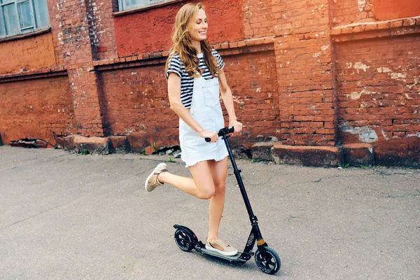 Giovane bella ragazza sta cavalcando uno scooter in estate lungo la strada sullo scooter e ride. Il concetto di uno stile di vita sano — Foto Stock