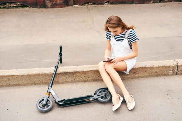 Genç güzel kız şehir ve görüşmeler bir scooter yanındaki telefonda yaz aylarında bir kaldırım kenarında oturuyor. Sağlıklı bir yaşam tarzı kavramı — Stok fotoğraf