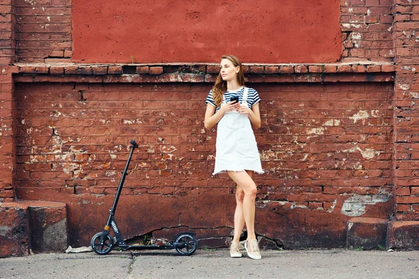 Joven chica hermosa se encuentra cerca de una pared de ladrillo rojo en verano en la ciudad y está hablando por teléfono al lado de un scooter. El concepto de un estilo de vida saludable — Foto de Stock