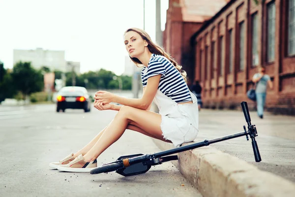 A menina bonita nova descansa e senta-se no passeio perto da estrada no verão na cidade ao lado de uma scooter. O conceito de um estilo de vida saudável — Fotografia de Stock