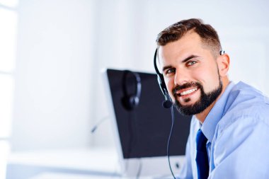 Yakışıklı destekçi gülümsüyor ve ofiste kulaklık ve bilgisayarla çalışıyor.