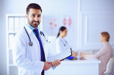 Hastanedeki gülümseyen Arap doktor tablet ve steteskopla arka planda hastayla birlikte çalışan tıbbi ekip