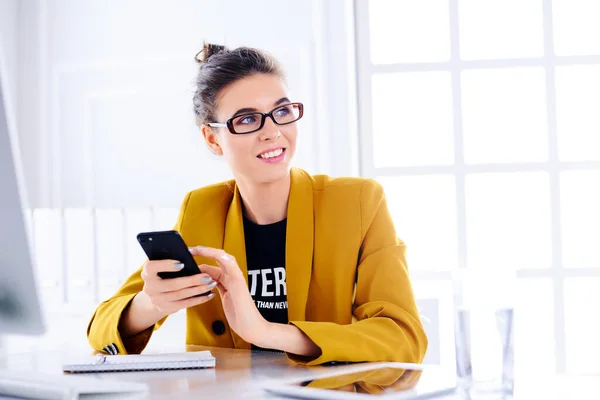 Business woman sits at a table in front of a computer in her office and holds a phone in her hands and smiling