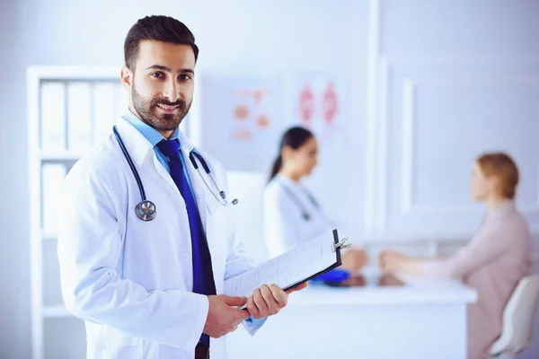 Hastanedeki gülümseyen Arap doktor tablet ve steteskopla arka planda hastayla birlikte çalışan tıbbi ekip
