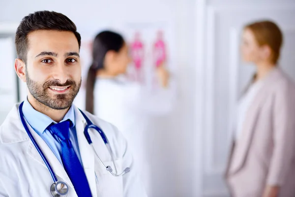 Ofisteki Arap doktor tablet ve steteskopla arka planda hastayla çalışan hemşire.