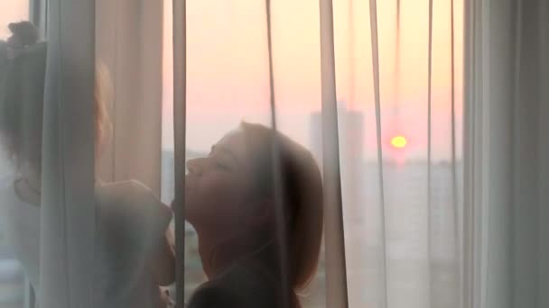 Η μητέρα και η κόρη της παίζουν κοντά στο παράθυρο μπροστά στο ηλιοβασίλεμα της πόλης. Οικογενειακή έννοια — Αρχείο Βίντεο