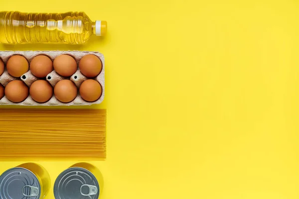 Eggs, pasta, canned food, oil in a paper bag on yellow background. Copy space, flat lay. Crisis food stock for coronavirus quarantine isolation period. Food delivery, donation.