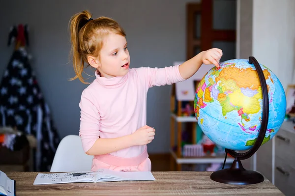 Young caucasian girl looking at earth globe at home. Schoolgirl doing homework and showing something at the globe