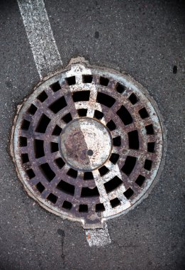 Üzerinde beyaz yol çizgileri olan asfalt kaplı metal kapaklı rögar..