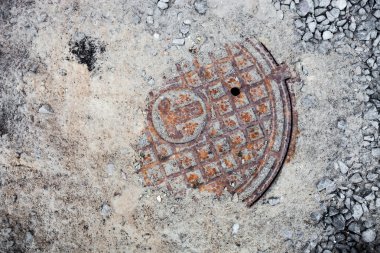 Manhole with the rusty cover partly in the sand and crushed stones clipart