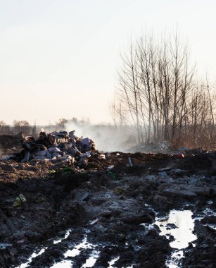 Mud and puddles on the dirt road. Burning garbage dump in the background. 