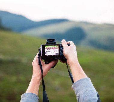 Dijital fotoğraf makinesini tutan eller. Kamera önizlemesine bakıyorum. Çalma ekranını kontrol ediyorum. Arkaplanda dağ manzarası.