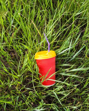 Sarı kapaklı ve mor kıvrımlı parlak kırmızı plastik bardak çimenlerde saman (tüp) içiyor.