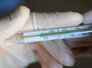 Ellerinde tıbbi eldivenler, camdaki tıbbi cıva termometresi var. Ateşi ölçüyorum. Yüksek ateş veya yüksek ateş, tehlikeli koronavirüs (Covid-19) enfeksiyonu da dahil olmak üzere birçok hastalığın belirtisidir.. 
