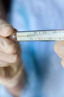 Ellerinde tıbbi eldivenler, camdaki tıbbi cıva termometresi var. Ateşi ölçüyorum. Yüksek ateş veya yüksek ateş, tehlikeli koronavirüs (Covid-19) enfeksiyonu da dahil olmak üzere birçok hastalığın belirtisidir.. 