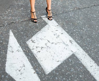 Yoldaki genç kadın. Yüksek topuklu sandaletli kadın bacakları. Yol çizgileri. Yoldaki beyaz oklar. 