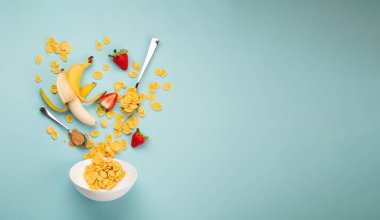 Sabahları hızlı ve sağlıklı bir kahvaltı: mısır gevreği, muz, beyaz kaseye taze çilek eklemek. Kahvaltını granola ile hazırlamak için malzemeler ve mavi arka planda meyveler, üst manzara, metin için boşluk