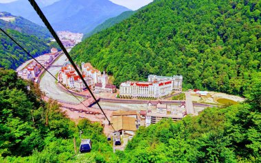 Sochi 'de, Rosa Khutor' da, yeşil ormanlı mavi gökyüzünün altında güneşli bir teleferik yolu.