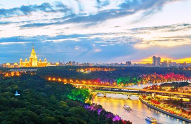 Günbatımının canlı panoramik manzarası. Köprüler, tekneler, güzel yansımalar ve ufuktaki ünlü Rus üniversitesinin ana binası ile aydınlatılmış Moskova nehri.