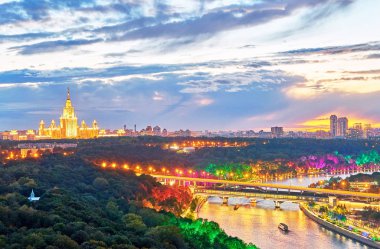 Günbatımının canlı panoramik manzarası. Köprüler, tekneler, güzel yansımalar ve ufuktaki ünlü Rus üniversitesinin ana binası ile aydınlatılmış Moskova nehri.