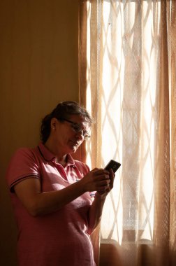 Pencerenin yanında cep telefonu olan yaşlı kadın telefonuna bakıyor. Karantina yüzünden akıllı telefonlu bir kadın evde bekliyor. Telefonunu kullanan bir bayan. Yalnız konsept. Karantina