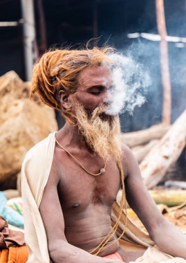 Prayagraj, Uttar Pradesh, India - February 2019: Indian naga sadhu smoking chilam cigar at biggest festival of Hindu - Kumbh Mela Fair. clipart
