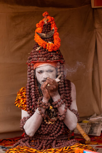 Prayagraj, Uttar Pradesh, India - February 2019: Indian naga sadhu smoking chilam cigar at biggest festival of Hindu - Kumbh Mela Fair.