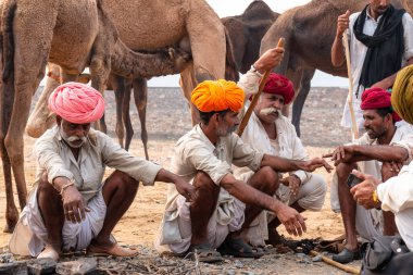 PUSHKAR, RAJASTHAN / INDIA - NOVEMBER 2019: Deve tüccarı / Hintli adamın etnik rajasthani elbisesi ve renkli sarıklı (Pagdi) deve panayırında develeri olan portresi