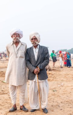 PUSHKAR, RAJASTHAN / INDIA - NOVEMBER 2019: Deve tüccarı / Hintli adamın etnik rajasthani elbisesi ve renkli sarıklı (Pagdi) deve panayırında develeri olan portresi