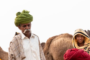 PUSHKAR, RAJASTHAN / INDIA - NOVEMBER 2019: Deve tüccarı / Hintli adamın etnik rajasthani elbisesi ve renkli sarıklı (Pagdi) deve panayırında develeri olan portresi
