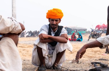 PUSHKAR, RAJASTHAN / INDIA - NOVEMBER 2019: Deve tüccarı / Hintli adamın etnik rajasthani elbisesi ve renkli sarıklı (Pagdi) deve panayırında develeri olan portresi