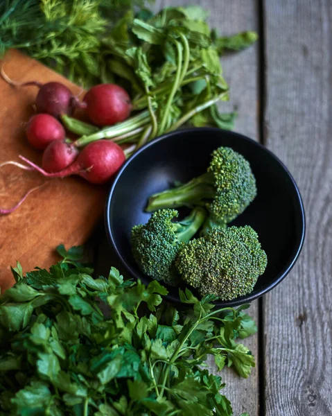 Broccoli in the bowl with radish and parsley and dill on the wooden board on grey wooden background prop. Top view.