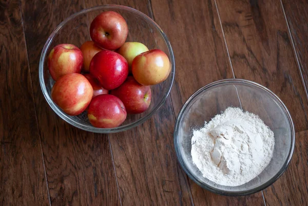 Clear glass bowl of apples and clear glass bowl of flour positioned near each other on a dark wood background