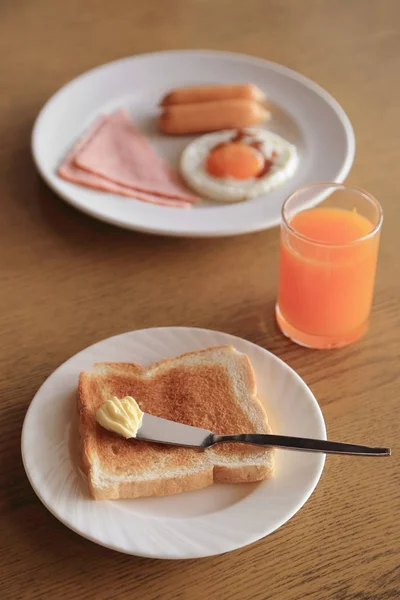 Conjunto de café da manhã, torrada com manteiga e suco de laranja, e prato de ovo frito, presunto, salsicha no fundo . — Fotografia de Stock