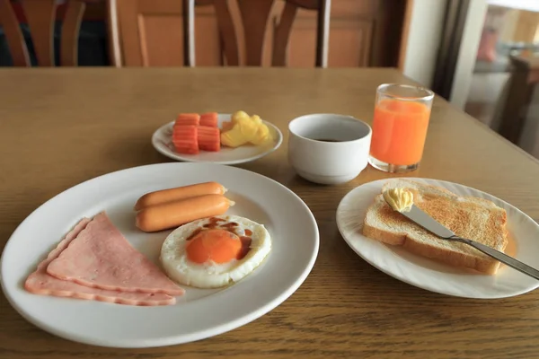 Conjunto de desayuno americano en mesa, huevo frito, jamón y salchichas, tostadas y mantequilla, frutas frescas, jugo de naranja y café negro . — Foto de Stock