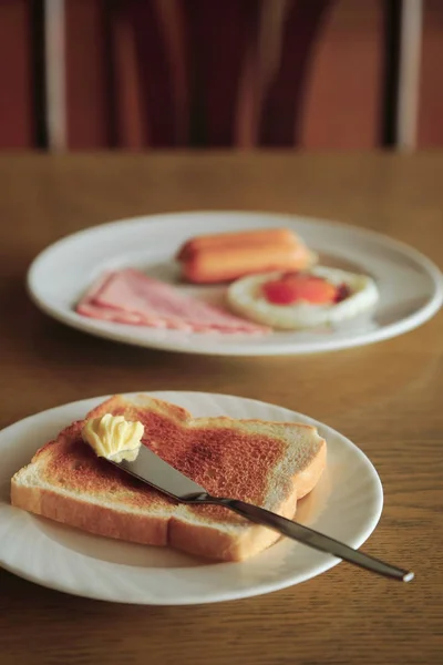 Café da manhã na mesa, torrada com manteiga e prato de ovo frito, presunto e salsicha no fundo, imagem vintage tonificada . — Fotografia de Stock