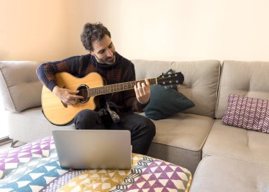 Evdeki kanepede oturan bir adam dizüstü bilgisayarla online gitar dersleri alıyor..
