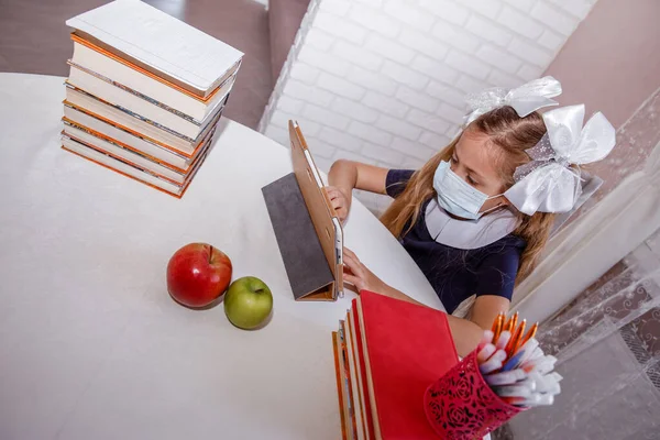 Distance learning online education. School girl in a medical mask is studying at home with a laptop and doing school homework. Textbooks and notebooks on the table