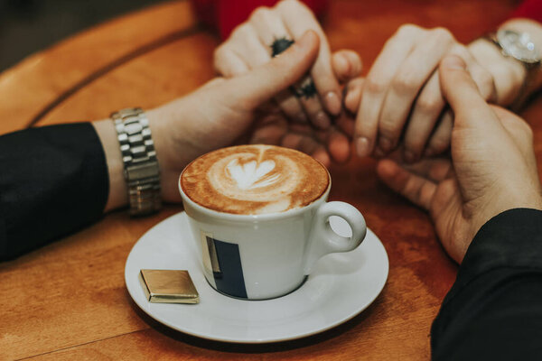 Руки мужчины держатся за руки девушки за чашку кофе на переднем плане на столе. Романтическая атмосфера. Закрыть
